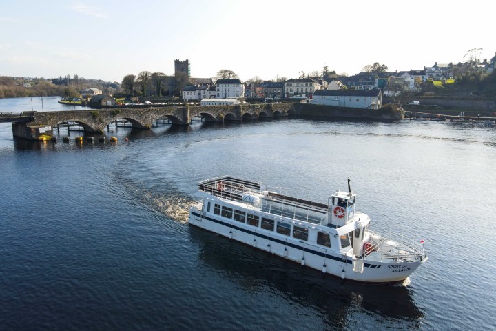 Passenger boat on a river near an arched bridge and town in daylight.