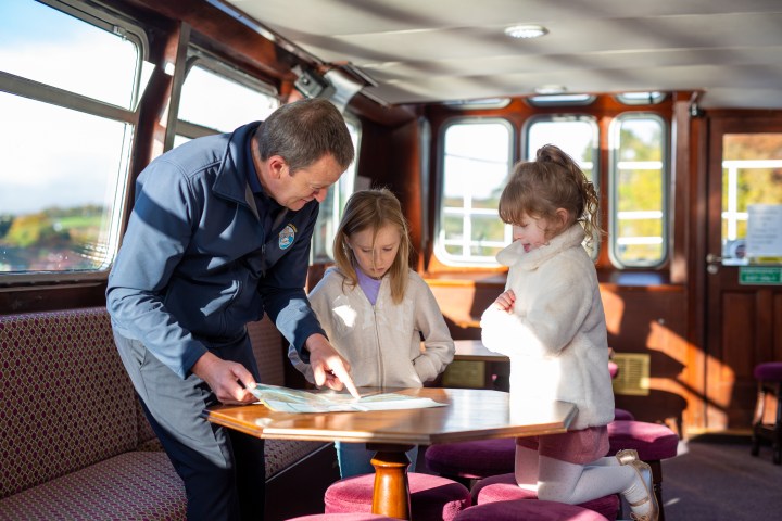 Man in uniform showing book to two young girls on a boat.