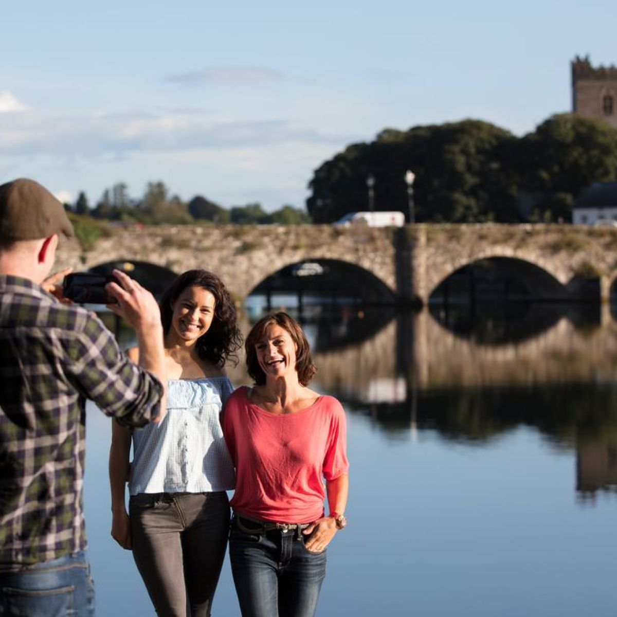 Person photographs two smiling people by river with stone bridge.