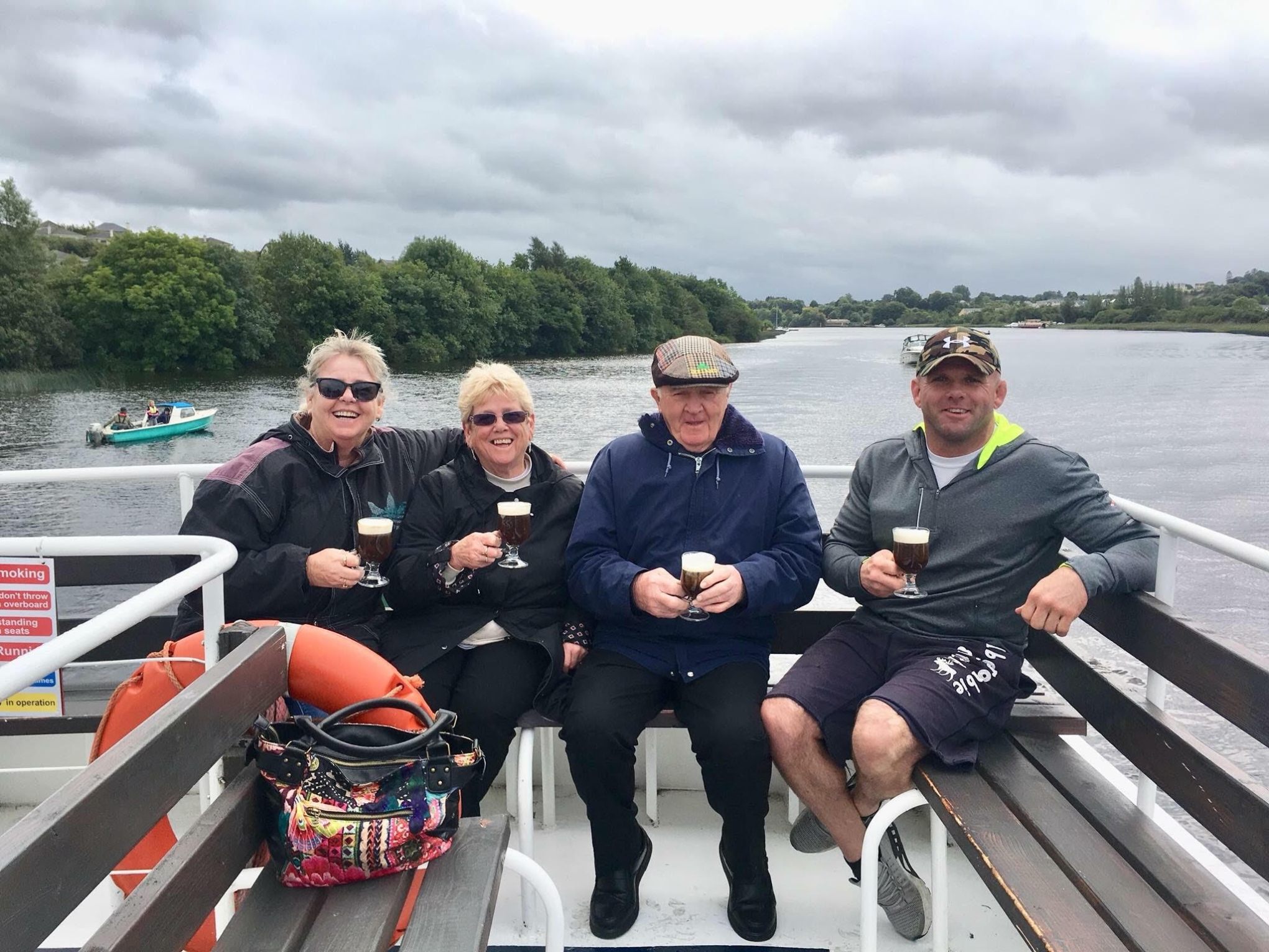 Four people smiling on a boat, holding drinks with trees and cloudy sky in the background.