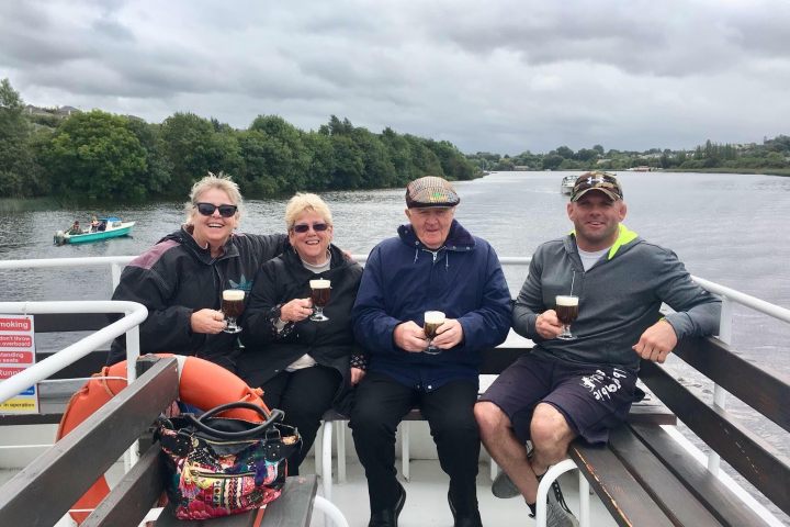 Four people smiling on a boat, holding drinks with trees and cloudy sky in the background.