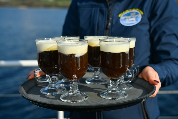 Person holding a tray of six Irish coffees in glass mugs on a boat.