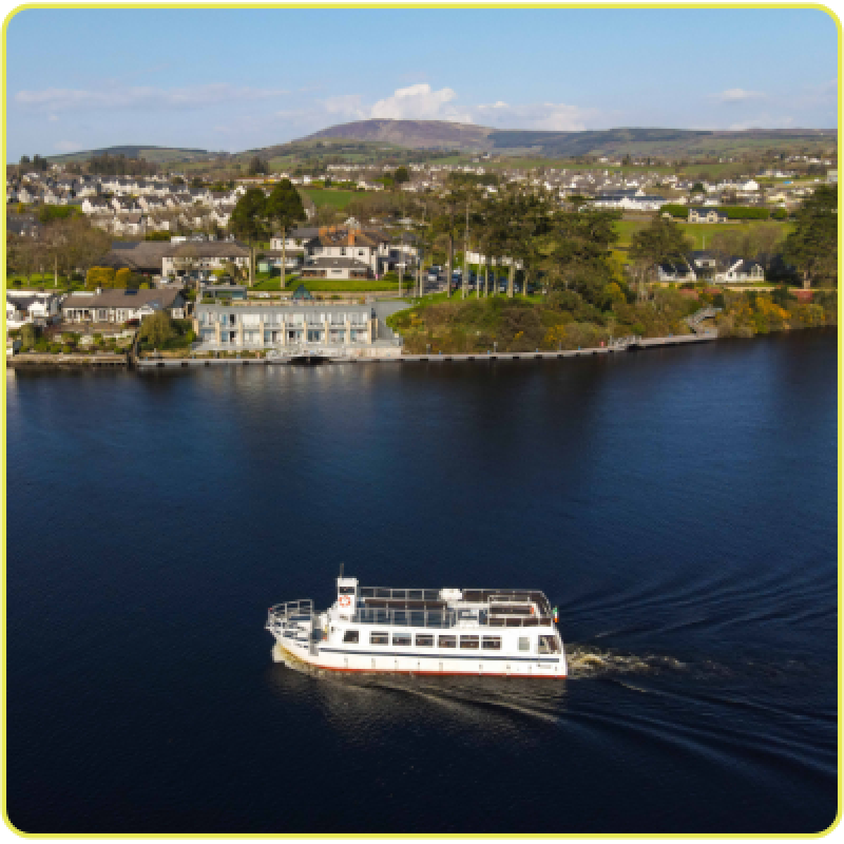 A white ferry boat on a calm river with a town and hills in the background.
