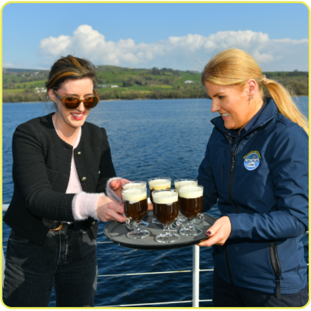 Two women on a boat holding Irish coffee glasses on a tray.