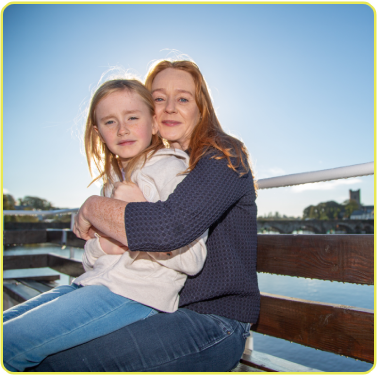 Woman hugging child on wooden bench with bright sun.