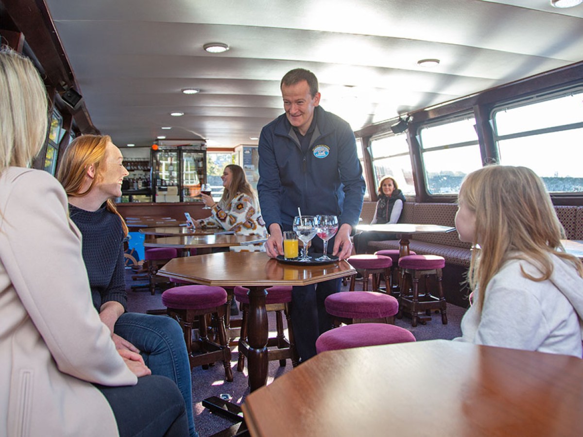 Server brings drinks to women and child on a boat with tables and seating.