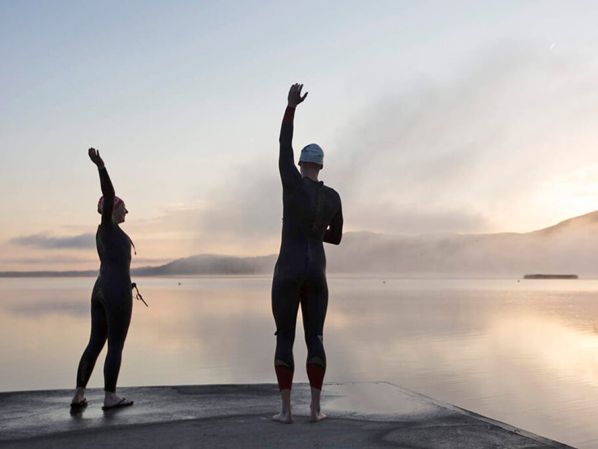 Two people in wetsuits wave by a calm lake at sunrise.