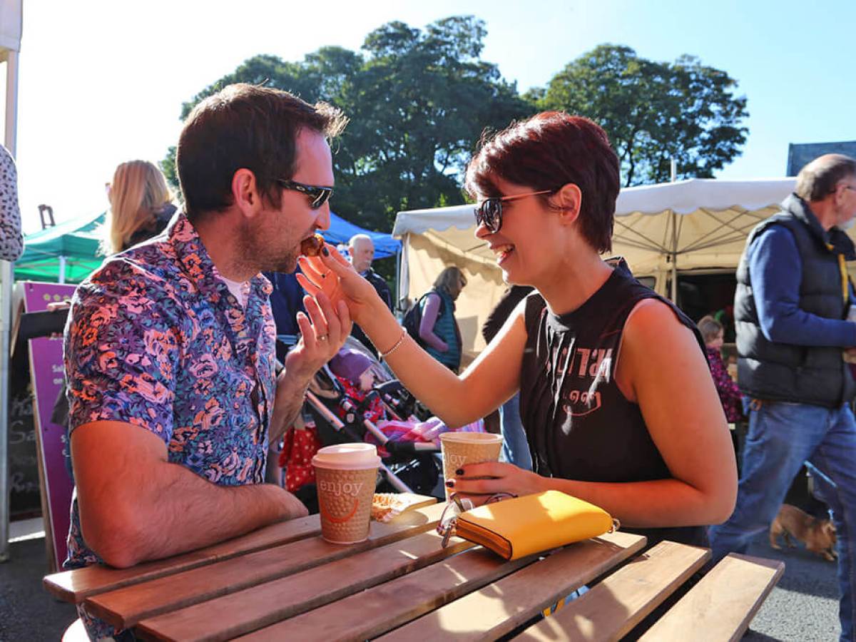 Couple laughing and sharing a drink at an outdoor market with trees in background.