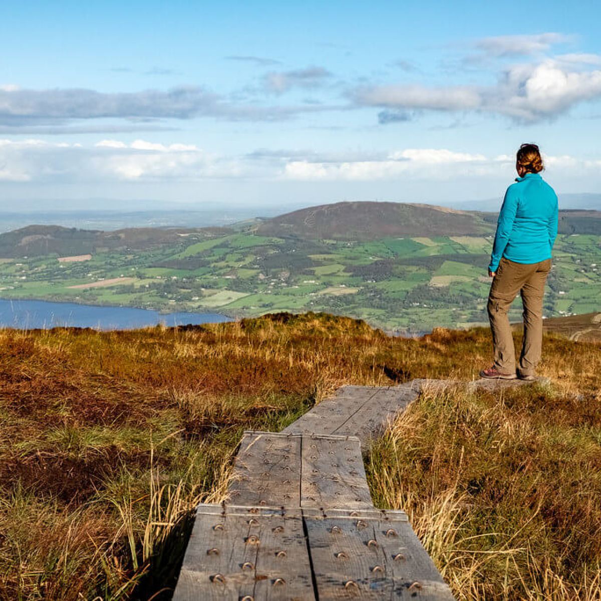 Person in blue jacket on wooden path overlooking hilly landscape.