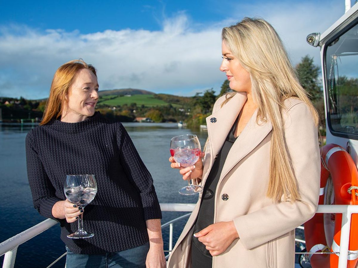 Two women on a boat holding drinks, with water and hills in the background.